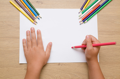 Pintura de la mano de la niña sobre el papel blanco con lápiz de color de rosa. Lápices de color sobre el escritorio de madera. Tiempo de dibujo. Vista superior. Lugar vacío. photo