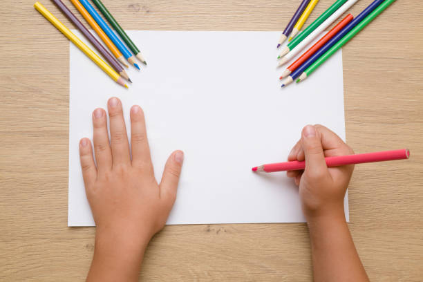 Little girl's hand painting on the white paper with pink pencil. Color pencils on the wooden desk. Drawing time. Top view. Empty place. Little girl's hand painting on the white paper with pink pencil. Color pencils on the wooden desk. Drawing time. Top view. Empty place. coloured pencil stock pictures, royalty-free photos & images