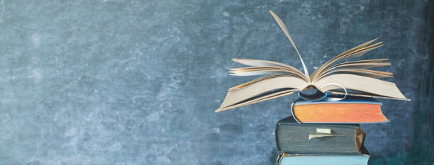 istock Open Book on a stack of old books in front of a blackboard, panoramic format, good copy space 1055093740