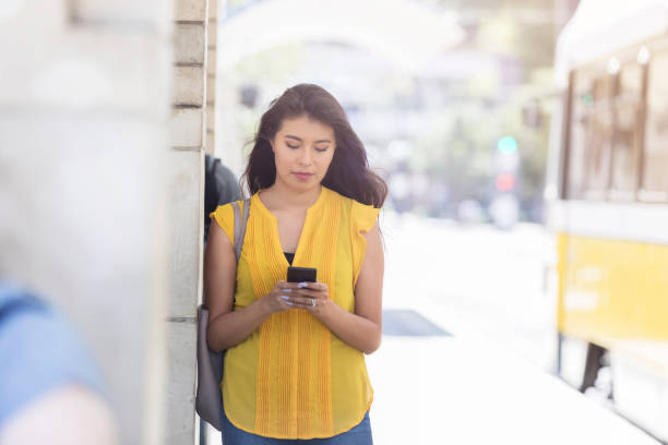 Young woman texts while waiting for bus A young woman leans against a column along a city sidewalk and texts on her smart phone as she waits for the bus. Distracted stock pictures, royalty-free photos & images