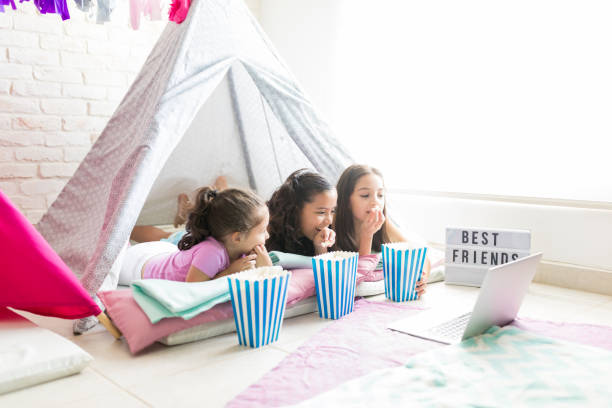 Female Friends Enjoying Snacks While Watching Laptop In Tent Female little friends enjoying snacks while watching film on laptop in tent during slumber party slumber party stock pictures, royalty-free photos & images