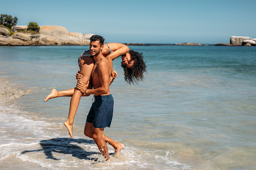 Man carrying his girlfriend on shoulder walking in the sea water on the shore. Couple enjoying and having fun at the sea.