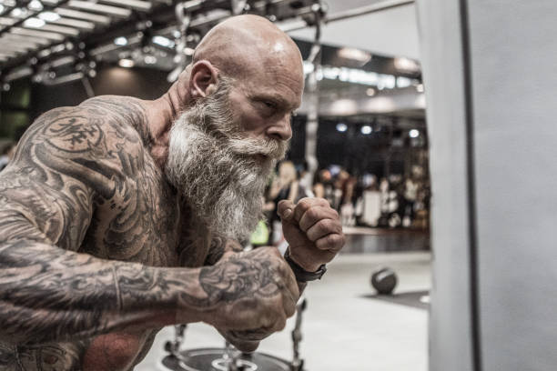 Tattooed Senior Man During Workout Bearded Aggressive Senior Man during workout in a public gym senior bodybuilders stock pictures, royalty-free photos & images