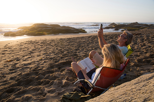 Mature woman uses tablet in beach chair as man photographs sunset, with digital tablet