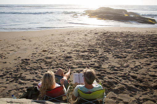 Mature couple sit in chairs on beach at sunset with tablet and book