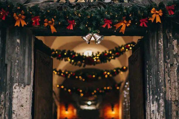 Photo of Stylish christmas decorations, jingle bells, garland lights, fir branches with ornaments on door front in european city street. Festive decor and illumination in city center, winter holidays