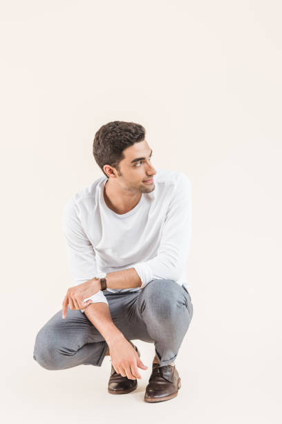 istock handsome smiling young man crouching and looking away isolated on beige 1055215454