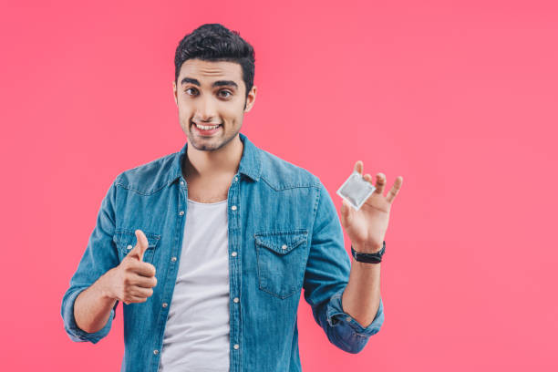 istock cheerful young man holding condom and doing thumb up gesture isolated on pink 1055216292