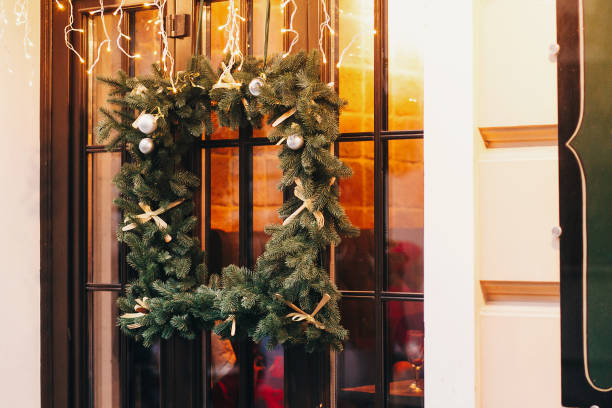 istock Stylish christmas square wreath with ornaments, pine cones, branches on window with lights in european city street. Festive decorations and illumination in city center, winter holidays. 1055217012
