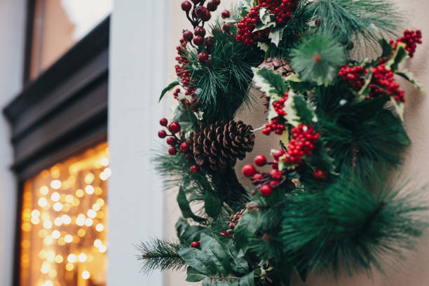 Stylish christmas wreath with red berries,ornaments, pine cones, branches on building in european city street. Festive decorations and illumination in city center, winter holidays.
