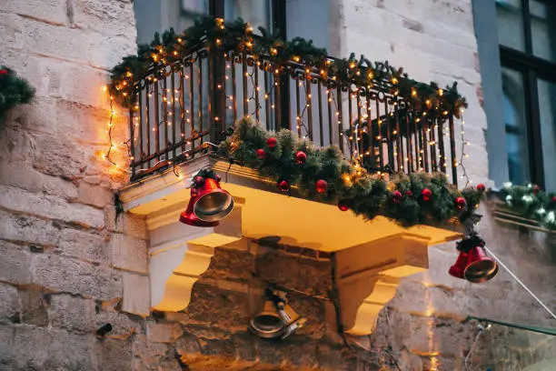 Photo of Stylish christmas decorations, red jingle bells, lights, fir branches with ornaments  on balcony  in european city street. Festive decor and illumination in city center, winter holidays