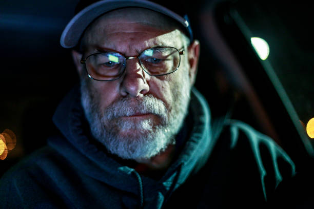 Senior adult man driving on a night road trip journey. He is in his car alone just after midnight. Light from an illuminated smart phone maps and navigation mobile app screen is reflected in his eyeglasses. It's winter in December and he's wearing a warm hoody-type hooded sweatshirt and a baseball cap as he concentrates on the directions for the road ahead.