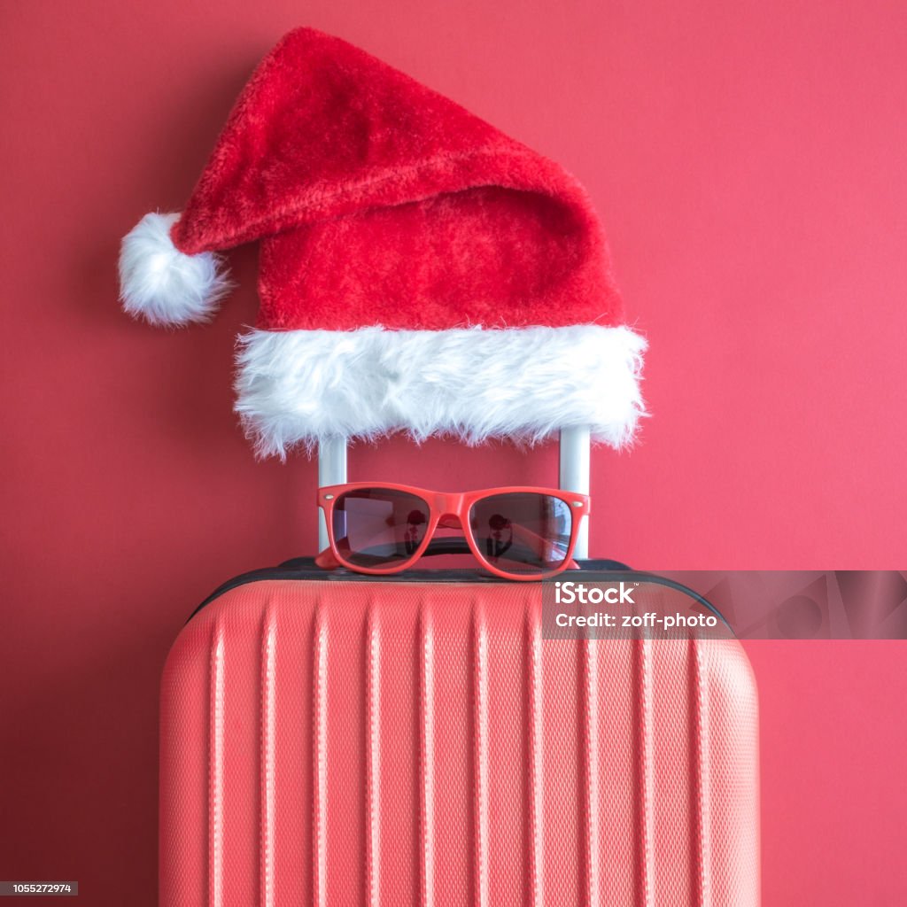 Flat lay of Santa Claus hat, sunglasses and luggage abstract isolated on red. Suitcase with Santa hat and sunglasses on red background minimal creative concept. Christmas Stock Photo