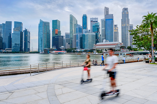 Modern skyline of Singapore with skyscrapers, people riding kick scooters at city embankment
