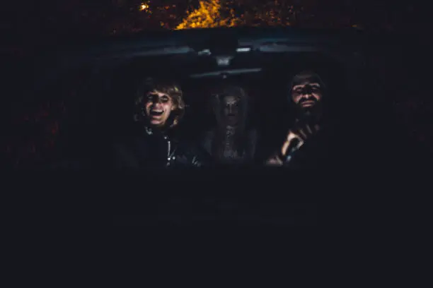 Man and woman sitting in a car, witch is sitting behind them, halloween night.