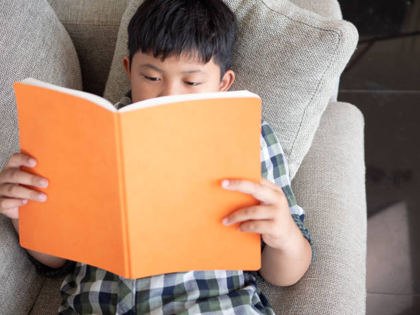 istock Cute asian children reading a book while lying on a sofa in the room. education concept 1055349810
