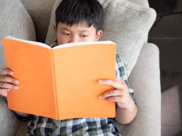 istock Cute asian children reading a book while lying on a sofa in the room. education concept 1055349816