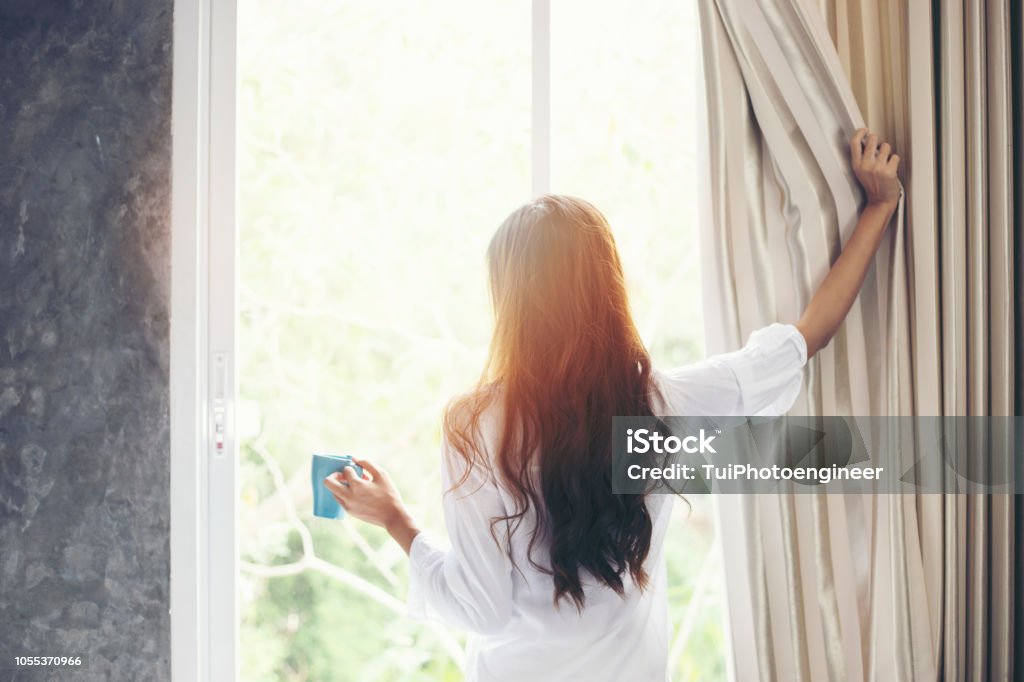 Asian women drinking coffee and wake up in her bed fully rested and open the curtains in the morning to get fresh air on sunshine Women Stock Photo