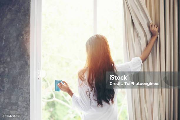 Asian Women Drinking Coffee And Wake Up In Her Bed Fully Rested And Open The Curtains In The Morning To Get Fresh Air On Sunshine Stock Photo - Download Image Now