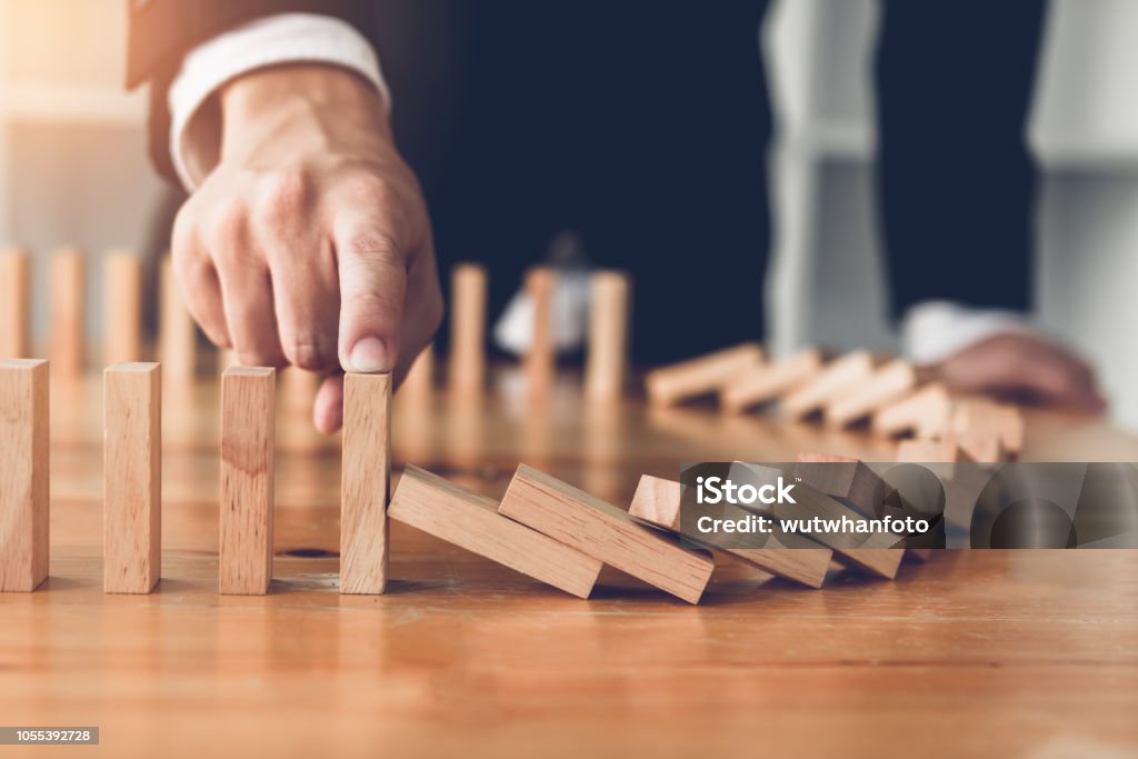 Close up finger businessman stopping wooden block from falling in the line of domino with risk concept. Risk Stock Photo