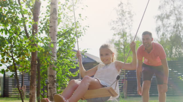 Joyous Father Pushes Swings with His Cute Little Daughter on Them . Happy Family Spends Time Together one Sunny Summer Day in the Idyllics Backyard.