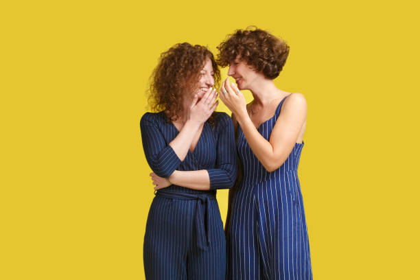 istock Handsome young caucasian student in dark blue overalls standing at yellow background, whispering something into ear of stylish ginger girl, sharing latest gossip. Woman looking in happy and smiling 1055420238