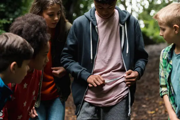 Young Boy showing off and pulling a knife out from underneath his jacket white his friends stand around in a woodland area.