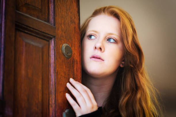 A beautiful young woman cautiously opens her front door, peeping round it nervously, worried about intruders.