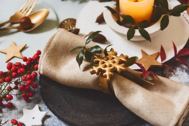 Holiday table setting, black plate with golden napkin holder with star shape, on rustic texture, surrounded by ornaments nandine branches and burning candles