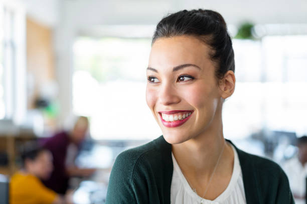 Close-up of beautiful young businesswoman standing at creative workplace. Smiling female professional is looking away at office. She is in smart casual.