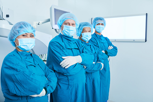 Side view of Asian surgeons in masks keeping arms folded and looking at camera while standing in modern operating theatre in hospital