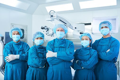 Asian surgical team in masks and uniform keeping arms crossed and looking at camera while standing in operating theatre together