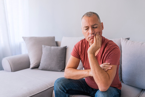 Photo of Depressed ill man having toothache and touching cheek. Mature man suffering from tooth pain, caries. Handsome gray hair male suffering from toothache, closeup. Portrait of casual 46 years mature man toothache with painful expression, sitting on sofa at home,