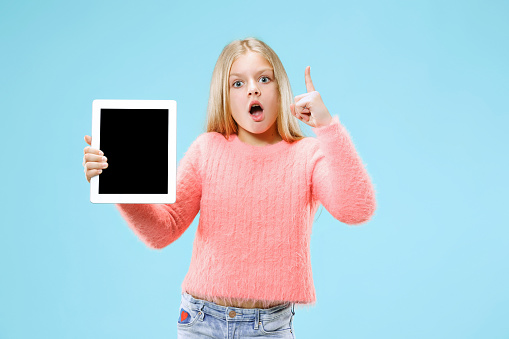 Little funny girl with tablet on blue studio background. She showing something and pointing up