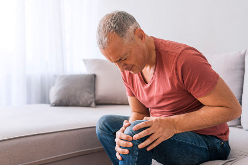 Photo of mature, elderly man sitting on a sofa in the living room at home and touching his knee by the pain during the day. Mature man massaging his painful knee.