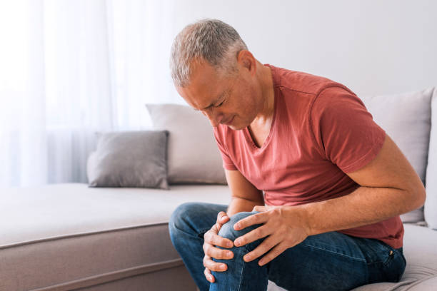 Feeling the aches of a long day Photo of mature, elderly man sitting on a sofa in the living room at home and touching his knee by the pain during the day. Mature man massaging his painful knee. knee photos stock pictures, royalty-free photos & images