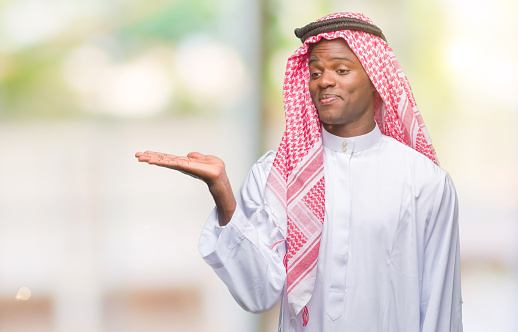 Young african man wearing traditional keffiyeh over isolated background smiling cheerful presenting and pointing with palm of hand looking at the camera.