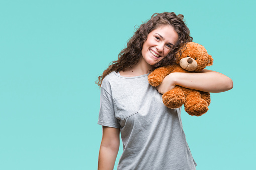 Young brunette girl holding teddy bear over isolated background with a happy face standing and smiling with a confident smile showing teeth