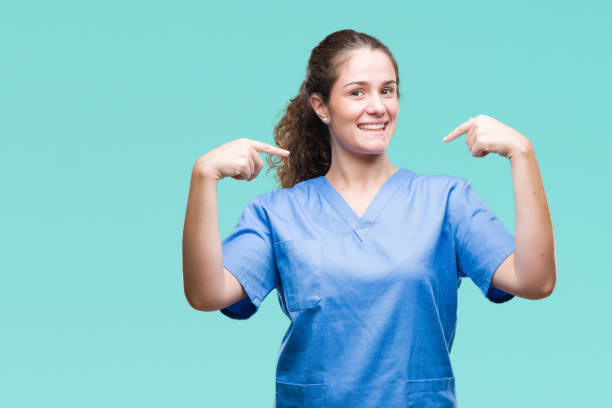 Young brunette doctor girl wearing nurse or surgeon uniform over isolated background looking confident with smile on face, pointing oneself with fingers proud and happy. Young brunette doctor girl wearing nurse or surgeon uniform over isolated background looking confident with smile on face, pointing oneself with fingers proud and happy. sign human hand pointing manual worker stock pictures, royalty-free photos & images
