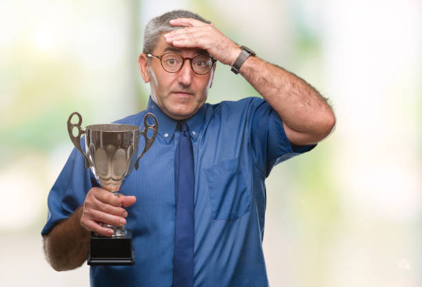 istock Handsome senior successful man holding trophy over isolated background stressed with hand on head, shocked with shame and surprise face, angry and frustrated. Fear and upset for mistake. 1055521124