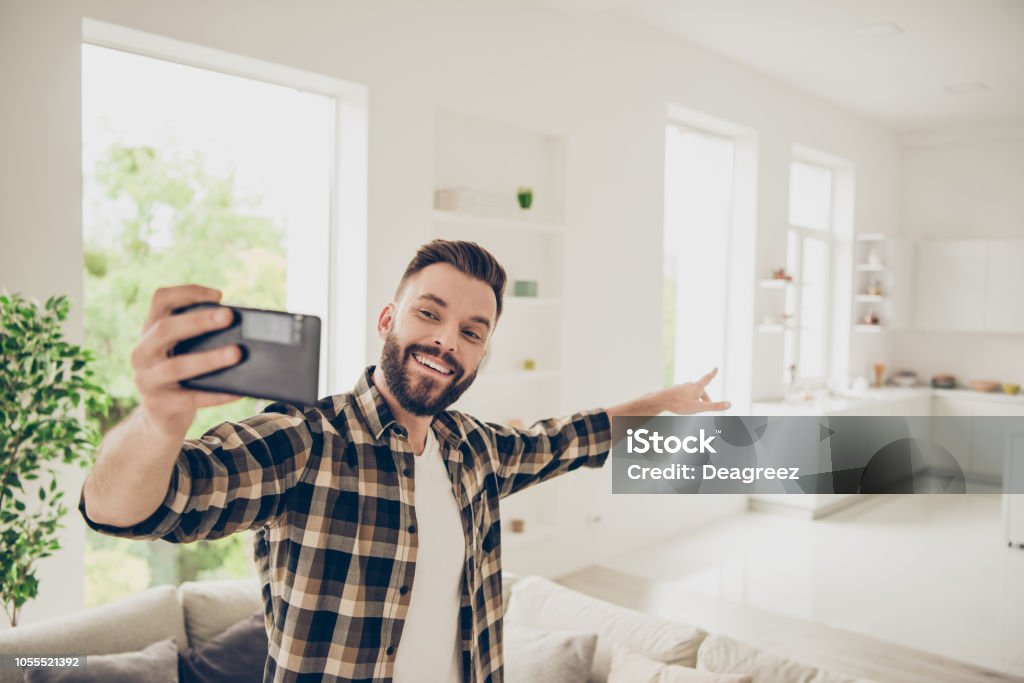 Apartment purchase concept. Let's go and I'll show you my house! Young man make call on front camera of modern smartphone standing in the middle of a light room in his brown stylish trendy shirt Selfie Stock Photo