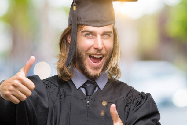 istock Young handsome graduated man with long hair over isolated background approving doing positive gesture with hand, thumbs up smiling and happy for success. Looking at the camera, winner gesture. 1055535016