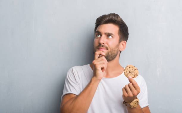 istock Handsome young man over grey grunge wall eating chocolate chip cooky serious face thinking about question, very confused idea 1055544858