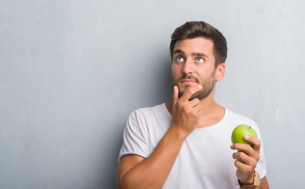 istock Handsome young man over grey grunge wall eating green apple serious face thinking about question, very confused idea 1055544860