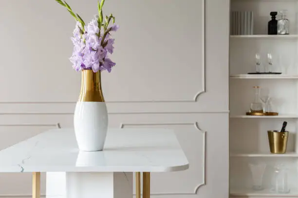 Photo of Lavender purple flowers in a golden vase on a white marble table in an elegant dining room interior with molding on beige walls