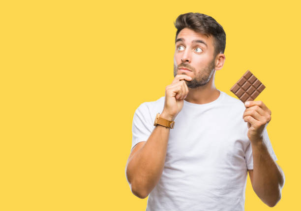 Young handsome man eating chocolate bar over isolated background serious face thinking about question, very confused idea