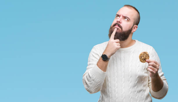 Young hipster man eating chocolate chips cookie over isolated background serious face thinking about question, very confused idea Young hipster man eating chocolate chips cookie over isolated background serious face thinking about question, very confused idea Smart Snacking stock pictures, royalty-free photos & images
