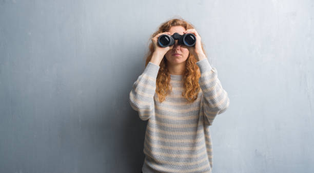 istock Young redhead woman over grey grunge wall looking through binoculars with a confident expression on smart face thinking serious 1055573012