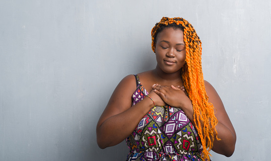 Young african american woman over grey grunge wall wearing orange braids smiling with hands on chest with closed eyes and grateful gesture on face. Health concept.