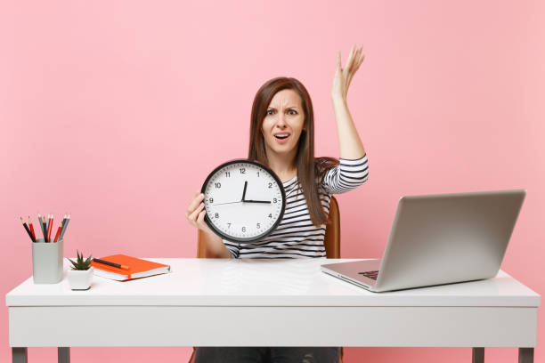 istock Young irritated woman spreading hands holding alarm clock sit, work at office with pc laptop isolated on pastel pink background. Achievement business career concept. Copy space. Time is running out. 1055594078